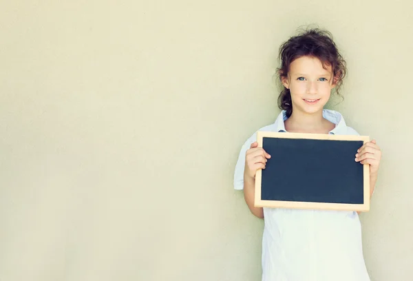 Miúdo bonito (menina) segurando quadro negro de pé contra a parede texturizada, imagem retro filtrada . — Fotografia de Stock