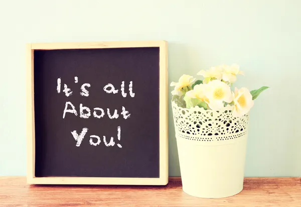 Blackboard with the phrase it's all about you written on it. over wooden shelf and flowers. — Stock Photo, Image