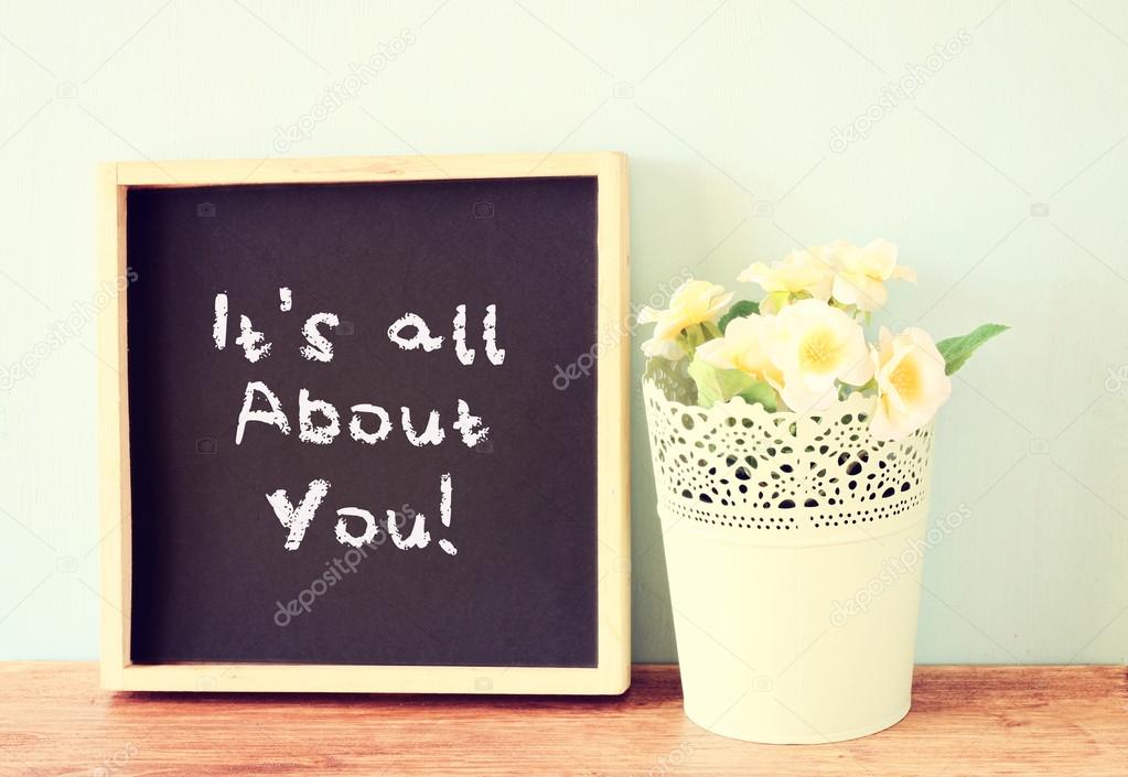 Blackboard with the phrase it's all about you written on it. over wooden shelf and flowers.