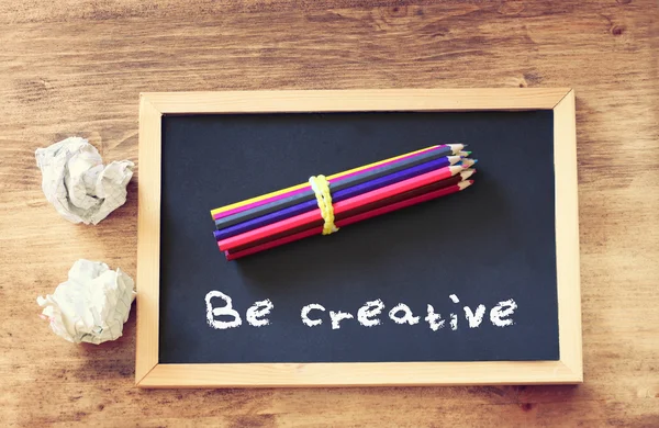 Top view of crumpled paper and pencils stack over blackboard with the phrase be creative. — Stock Photo, Image