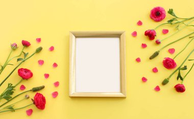 Top view image of pink buttercup flowers composition and empty gold photo frame over yellow pastel background