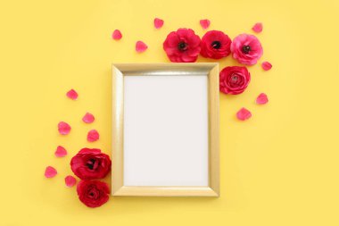 Top view image of pink buttercup flowers composition and empty gold photo frame over yellow pastel background