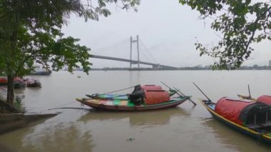 Arka planda ikinci Howrah Köprüsü 'nün yanındaki Ganj Nehri kıyısında eğlence amaçlı tekneler var. Ön plandaki ağaçları çerçevelemek. Yağmurlu bir gün. Kolkata Babu Ghat Hindistan Güney Asya Pasifik. 16 Ekim 2021
