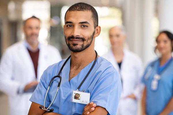 面带微笑的中东男人身着制服 用听诊器看着相机的画像 年轻医生笑着站在医院走廊里 背后有医护人员 成功的印度外科医生在他的多种族医学面前微笑 — 图库照片