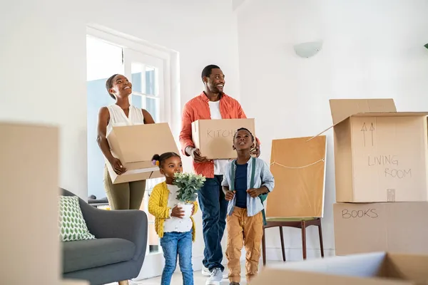 Beautiful african american family with two children carrying boxes in a new home. Cheerful mature mother and mid adult father holding boxes while entering new home with son and daughter. Happy son and daughter helping parents relocating in new house