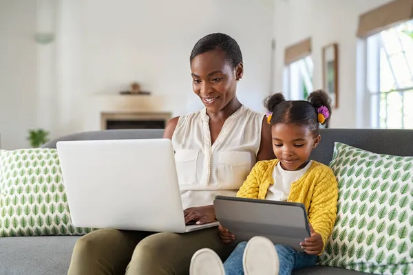 在家里用笔记本电脑微笑成熟的黑人妈妈 而在沙发上坐在旁边的可爱小女孩用数码平板电脑 快乐的非洲成熟女人在家里用笔记本电脑远程工作 帮助女儿做作业 自由撰稿人中老年沃马 — 图库照片