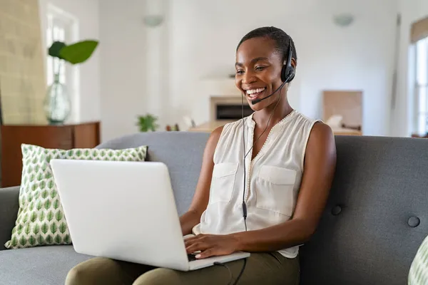 成功的中年非洲裔美国女人 带着耳机和麦克风 用笔记本电脑通过视频通话 快乐的中年黑人妇女坐在沙发上 一边坐在笔记本电脑上一边看电视一边聊天 快乐微笑的女士一边用电脑进行视频会议一边 — 图库照片