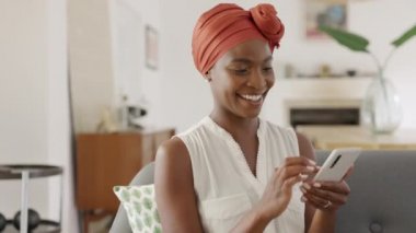 Geleneksel türbanlı gülümseyen orta yaşlı Afrikalı kadın akıllı telefon kullanarak evdeki kanepede oturuyor. Tipik türbanlı güzel Afro-Amerikan kadın cep telefonuyla internette geziniyor. Mutlu orta yaşlı bayan cep telefonuna bakıyor..