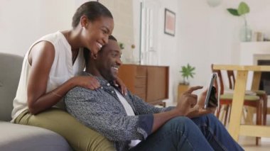 İnternette sörf yapmak için kanepeye otururken orta yaşlı siyah çift dijital tablet kullanıyor. Mutlu gülümseyen kadın dijital tablette video izlerken erkek arkadaşını kucaklıyor. Afrikalı kadın evde kocasıyla video görüşmesi yapıyor..
