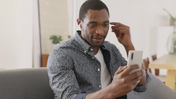 スマートフォンを使ってアフリカの成人男性の笑顔でビデオ会議を行います 友人とビデオ会議で話している間 中年の笑顔の男は自宅で休んで 無線イヤフォンを使用しています 自宅でビデオチャットを使用しているビジネスマン — ストック動画