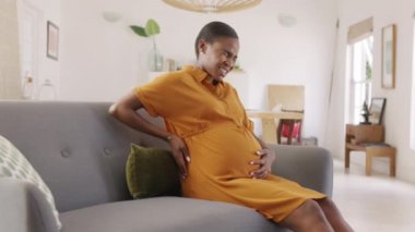 Olgun hamile bir kadın evdeki koltukta dinlenirken sırt ağrısı çekiyor. Mutsuz siyahi kadın bel ağrısından muzdarip. Orta yaşlı, Afrikalı hamile bir kadın geçen ayki hamileliğinde bel ağrısından muzdarip..