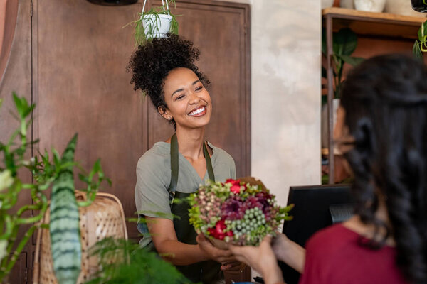 Portrait of smiling black botanist selling a bouquet of fresh flowers to a customer in a botany shop. Happy african woman entrepreneur standing behind counter wearing apron and selling bunch of fresh flowers to client. Young latin woman buying plant 