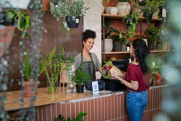 Smiling african woman botanist selling flowers and plants to a customer while standing in flower shop. Happy black young woman entrepreneur standing behind counter wearing apron in plant store selling fresh flowers to client. Young latin girl buy a f