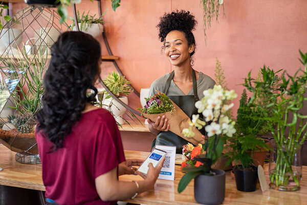 Young african florist holding beautiful bouquet of fresh flower while customer making online payment with smartphone while scanning QR code. Black saleswoman wearing apron giving a bunch of flower to customer at botany shop. Young woman making digita
