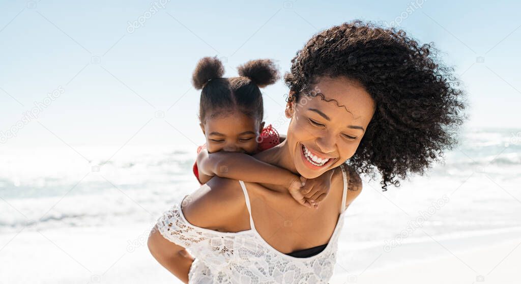 Happy black young woman giving piggyback ride to her daughter at beach with copy space while laughing. Cute african little girl with bigger sister playing outdoor at seaside. Close up of beautiful and carefree mom carrying on shoulders her funny fema