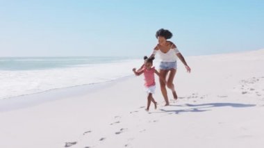 Beyaz tropikal plajda çıplak ayakla koşan küçük tatlı bir kızla mutlu siyah bir anne. Güzel, kıvırcık saçlı kız kardeşiyle kumsalda koşan neşeli Afrikalı Amerikalı kadın. Gülümseyen anne ve komik kadın çocuk denizde eğleniyor..