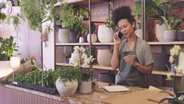 식물학 가게에서 쓰면서 스마트폰으로 이야기하는 미소짓는 판매원 아프리카 미국인 식물학자는 — 비디오