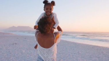 Sahilde kızının etrafında dönen ve sarılan neşeli genç bir anne. Genç anne gün batımında siyah kızını sallarken eğleniyor. Anne ve kızı, altın ışıkta, yaz kumsalında aşkla oynuyorlar..