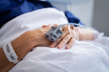 Hastanedeki hastanın elini serum fizyolojik solüsyon tüpüyle kapatın. Damardan serum verin. Yaşlı bir kadının kırışık ellerine yakın çekim. Hastaneye kaldırıldı ve özel klinikte tedavi altına alındı. Kan oksijeni saturasını ölçmek için indeksteki nabız oksimetrelerinin ayrıntıları