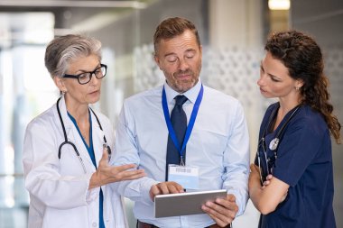 Kıdemli kadın doktor hastane koridorunda tıbbi personelle konuşuyor. Doktor ameliyattan sonra ekibiyle hasta durumunu tartışıyor. İlaç temsilcisi dijital tablet hakkında yeni ilaç ya da terapi raporu gösteriyor.