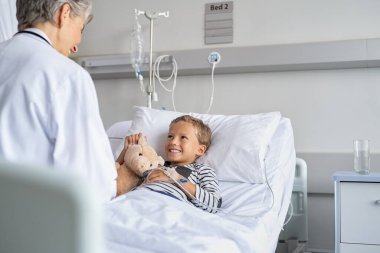 Neşeli olgun kadın doktor elinde damlalarla hastane yatağında yatan küçük çocuğu ziyaret ediyor. Çocuk doktoru onu ziyaret ederken oyuncak ayısını kucaklayan gülümseyen bir çocuk. Oksijen dolu sondası olan yakışıklı çocuk doktorla konuşuyor. 