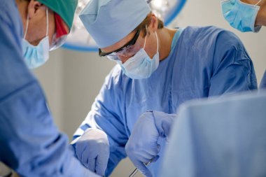 Deneyimli doktorlardan oluşan bir ekip ameliyathanede hastaya ameliyat yapıyor. Ameliyathanede ameliyat maskesi takan profesyonel bir cerrah makası tutuyor. Konsantre cerrah ameliyathanede hasta üzerinde çalışıyor.