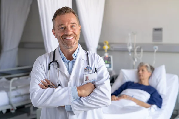 快乐成熟的医生 身穿带有听诊器的工作服 交叉双臂看着相机 在病人休息的时候 微笑着全科医生站在病床上 在私人诊所工作的成功医生的画像 复制空间 — 图库照片