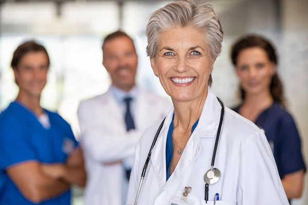 带着微笑的女医生与背景的团队站在医院里的画像 成熟的女医生在医护人员面前看着镜头 医院医务专业人员和医务工作者小组 — 图库照片