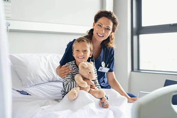Portrait of cheerful little boy holding teddy bear while sitting with beautiful nurse on gurney at hospital. Happy female nurse in pediatric ward with kid looking at camera. Young pediatrician doctor visiting and comforting hospitalized child at priv