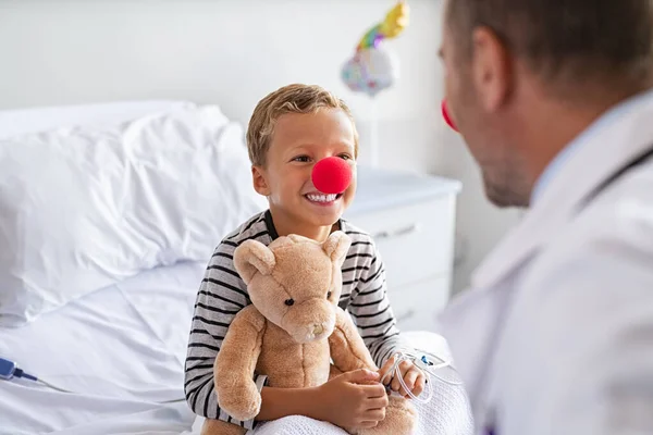 生病的小男孩头戴红鼻子 坐在病床上与医生玩耍 同时抱着泰迪熊 在诊所里与儿科医生交谈的小丑鼻子快乐孩子的画像 与全科医生一起住院的快乐儿童 — 图库照片