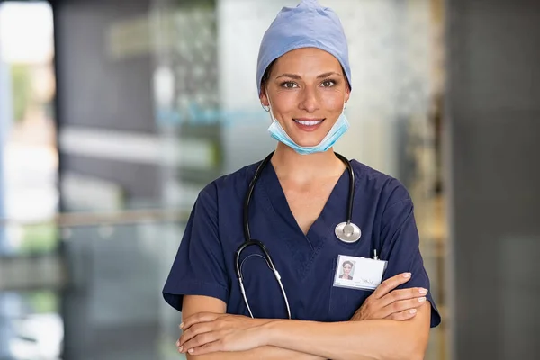 年轻女医生头戴口罩 在医院里擦拭的形象 自信可靠的医生 带着外科口罩 看着相机 微笑的护士 双手交叉 站在门诊部的走廊上 复印空间 — 图库照片