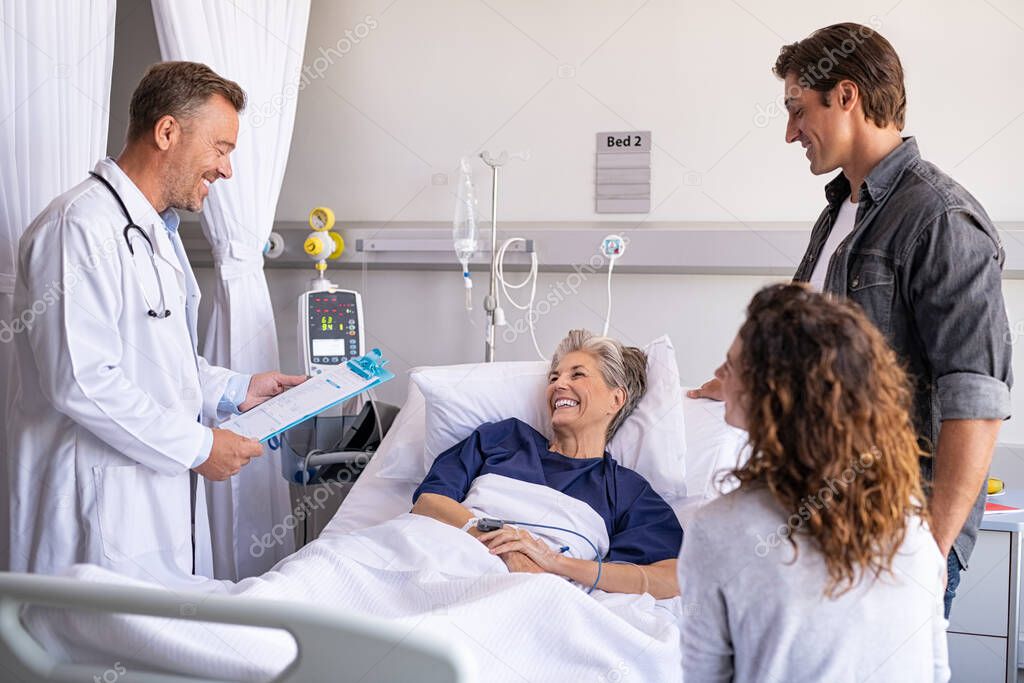 Mature happy doctor holding clipboard giving good news to family and old patient. Smiling senior woman lying on hospital bed with son and daughter visiting and talking to physician. Doctor visiting hospitalized patient with family in modern hospital 