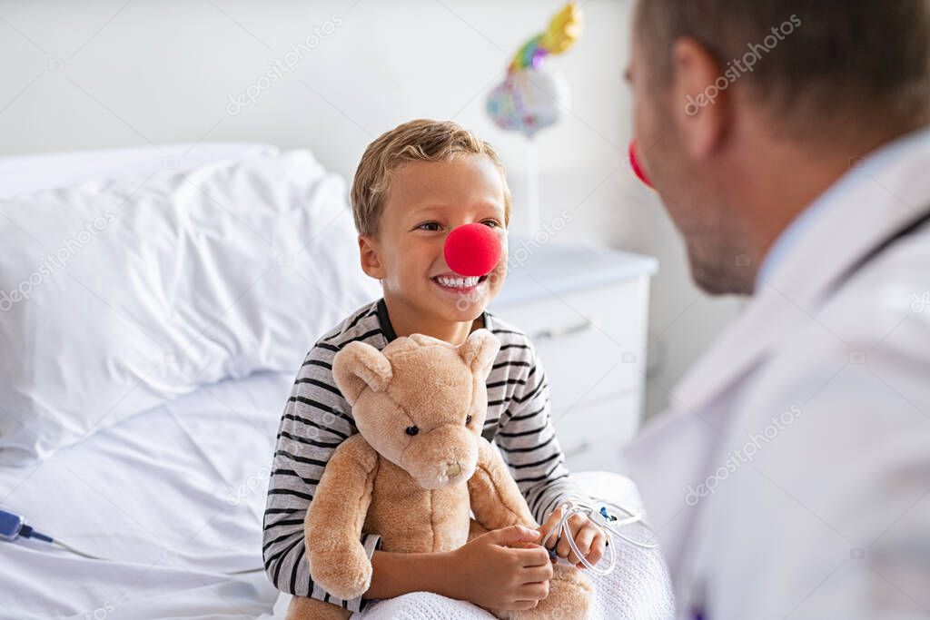 Sick little boy wearing red nose sitting on hospital bed playing with doctor while holding teddy bear. Portrait of cheerful kid with clown nose talking to pediatrician at clinic. Happy hospitalized child in hospital with general practitioner during a