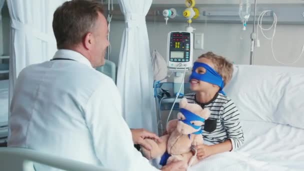 医者と遊んでいる間にテディベアを抱きかかえ病院のベッドに座っているスーパーヒーローマスクを持つかわいい男の子 小児科医と話をするおもちゃを持ったスーパーヒーローの格好をした遊び心のある子供 成熟した医者は入院中の彼の小さな患者を訪問 — ストック動画