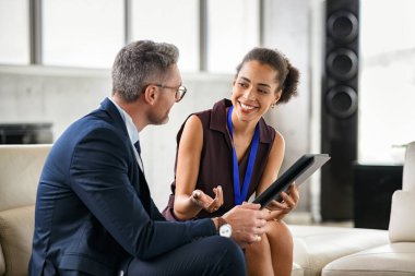 Young black businesswoman discussing work in office using digital tablet. African american woman manager in conversation with executive during conference. Professional mature businessman and smiling happy woman working while sitting on couch and disc