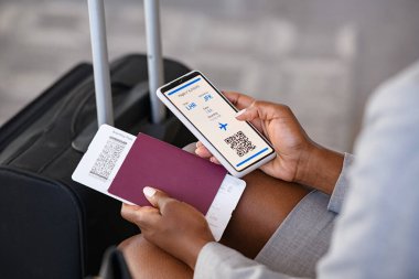 Close up hand of black businesswoman checking flight eticket on phone while holding passport and boarding pass. African american woman hand checking online travel plane ticket on mobile phone. Detail of business woman with flight ticket in waiting ro