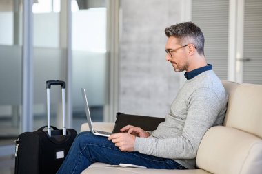 Successful mature man working on computer at airport in waiting room. Mid adult business man with luggage in international airport working on laptop while waiting for flight. Casual businessman passenger sitting on sofa in terminal hall and send an e