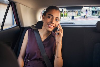Young smiling african woman talking over phone while sitting in taxi with copy space. Happy businesswoman sitting on back seat of car while using mobile phone to communicate with her boss. Successful woman talking over cellphone while commuting.