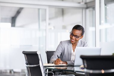 Mature businesswoman taking notes in notebook while using laptop at office. Mid adult black woman entrepreneur writing details on book while working on laptop in modern office. Smiling african american business woman with spectacles writing schedule 