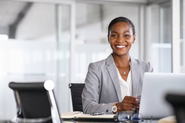 Portrait of a cheerful businesswoman sitting at desk in modern office and looking at camera. Attractive and successful black business woman sitting at conference table with laptop. Smiling african american executive using laptop while working from of