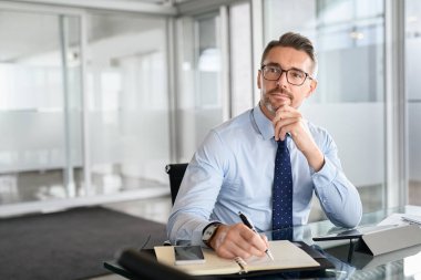 Thoughtful mature business man taking notes in notebook while using digital tablet at office. Mid adult businessman entrepreneur writing details on book while working in modern office. Pensive businessman with spectacles planning with copy space.