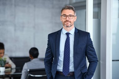 Portrait of handsome mid adult business man standing in modern office. Successful mature entrepreneur in formal clothing looking at camera with satisfaction. Confident man in suit with eyeglasses and beard standing with hands in pocket and looking at