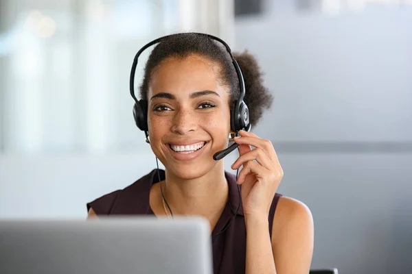 Call center agent with headset working on support hotline in modern office. Young positive african american agent in conversation with customer over headset looking at camera. Portrait of black girl working on phone call with laptop.