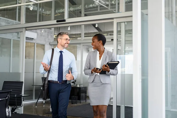 Smiling mature business man and african american businesswoman walking out of office. Two happy multiethnic colleagues talking while going home after work. Successful business people talking to each other in modern office with copy space.