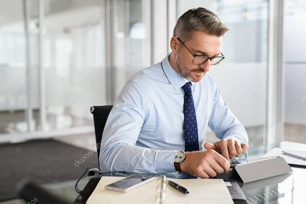 Busy businessman using digital tablet at work. Focused mid adult man working on computer in a modern office. Middle aged business man sitting at desk and checking email with copy space.