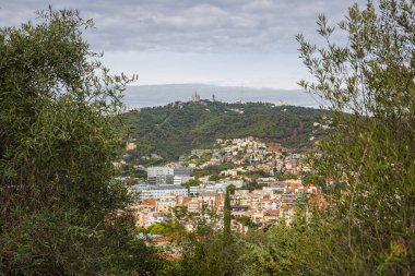 Barselona, İspanya - 22 Eylül 2021: Castell de Montjuic 'ten Tibidabo Dağı' ndaki İsa 'nın Kutsal Kalbi Kilisesi' ne bakın. Ağaçların arasından katedrale giden şehir manzarası.