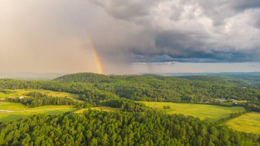 Kanada manzarasının uçsuz bucaksız ufkunda gökkuşağı. Yeşil çayır, tepedeki yemyeşil orman. Mavi gökyüzüne karşı yağmur bulutları, kara bulutların arasında güneş ışınları parlıyor. Ufukta gökkuşağı beliriyor