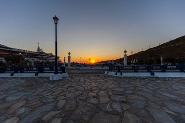Yeni Port Mykonos 'ta günbatımı. Limanın rıhtımında büyük bir yolcu gemisi var. Sarı kırmızı güneş ufukta Akdeniz 'e batıyor. Kaldırımdaki eski sokak lambaları..