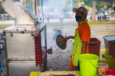 Kuala Lumpur, Malaysia - June 21, 2022: Young man barbecue chicken wings on open fire. He keeps the fire burning with a frond and smoke rises from the stove. On a street food market in the Malaysia