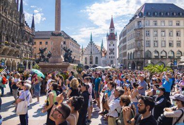 Münih, Almanya - 6 Temmuz 2022: Marienplatz 'daki turistler yeni belediye binasındaki saat zillerine hayret ediyorlar. Ziyaretçilerin kafalarının üzerinden eski belediye binasına ve Viktualienmarkt geçidine bakın..